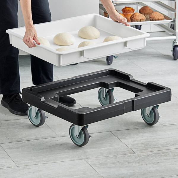 A woman using a black plastic dough box dolly to transport a white container of dough.