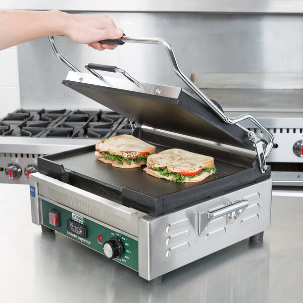 A hand placing a sandwich with lettuce and tomato on a Waring panini grill.