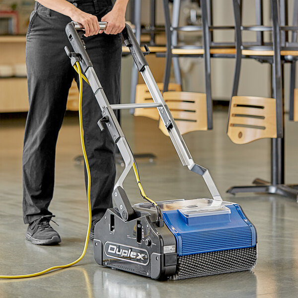A woman using a NaceCare Solutions Duplex walk behind floor scrubber to clean a floor.