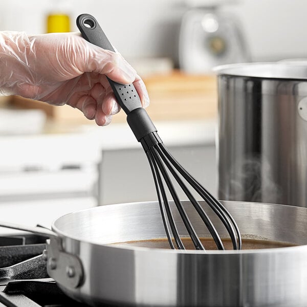 A hand holding a black Choice 12" heat-resistant nylon piano whisk over a pot of liquid.