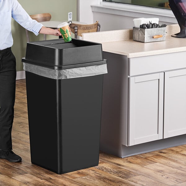 A man standing next to a Lavex black square trash can with swing lid.