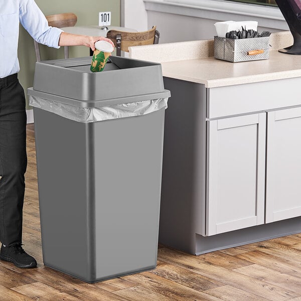 A person standing next to a Lavex 50 gallon grey square trash can with a swing lid.