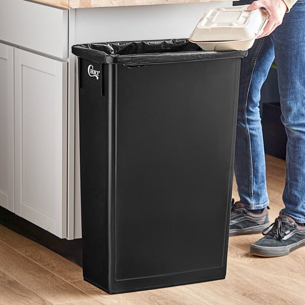 A person in blue jeans using a black rectangular Choice trash can with a white handle to throw away trash in a kitchen.