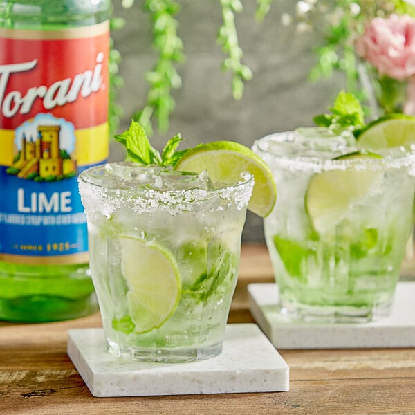 Two glasses of limeade with lime slices on a table with a bottle of Torani Lime Flavoring Syrup.