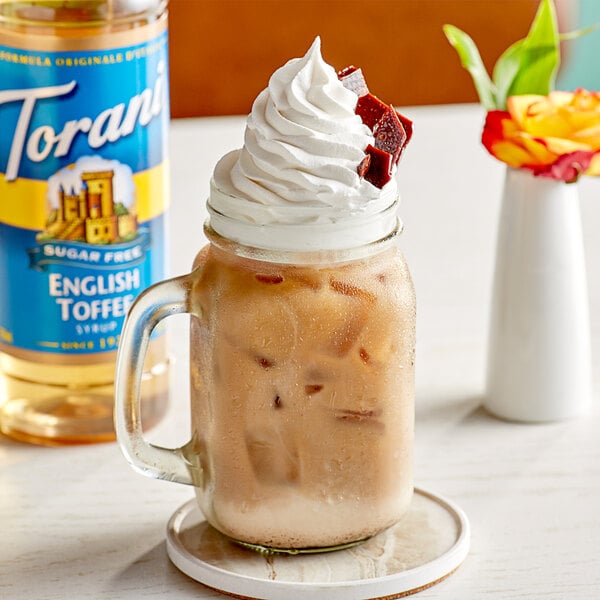 A glass mug with a drink and whipped cream on top with a bottle of Torani Sugar-Free English Toffee Flavoring Syrup.