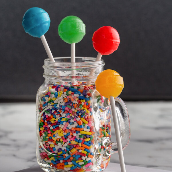 A glass jar filled with colorful lollipops and sprinkles on a counter with a red and blue lollipop on a Paper Lollipop Stick.