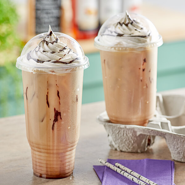 Two Choice clear plastic cups of iced coffee with whipped cream and chocolate syrup with dome lids.
