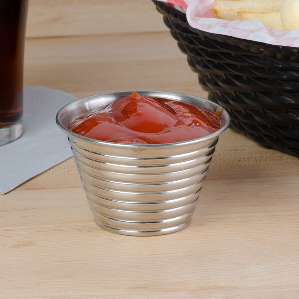 An American Metalcraft stainless steel sauce cup filled with ketchup on a table with a basket of fries.