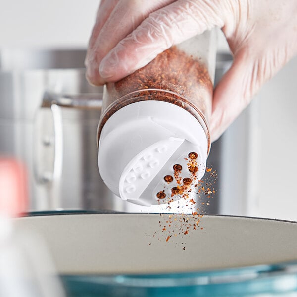 A hand with a red spice shaker pouring seasoning into a container with a white dual-flapper lid.