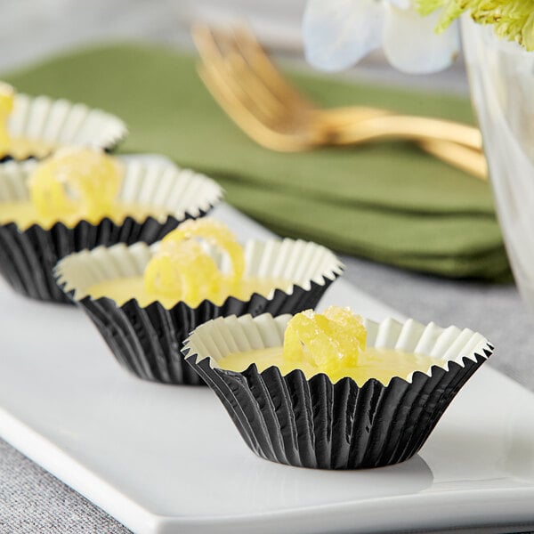 A row of Enjay black foil mini baking cups filled with lemon curd on a white plate.
