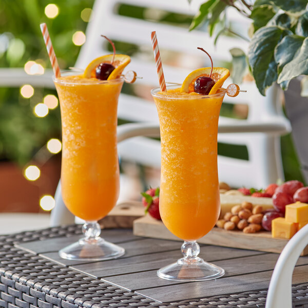 Two Acopa Endure Tritan plastic hurricane glasses of orange juice with straws and fruit slices on a table.
