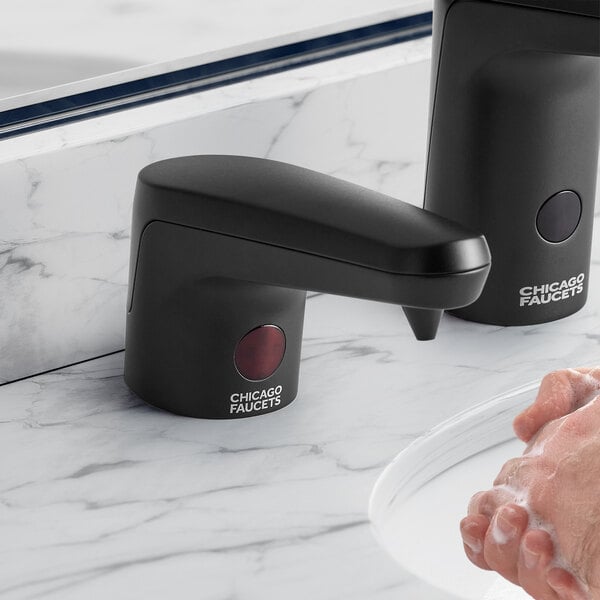 A person using a Chicago Faucets matte black touchless liquid soap dispenser on a counter.