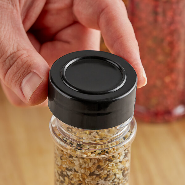 A hand holding a black round 43/485 polypropylene spice cap over a jar of seeds.