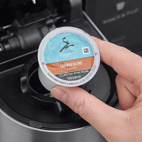 A hand holding a Caribou Coffee Caribou Blend K-Cup pod over a coffee cup.
