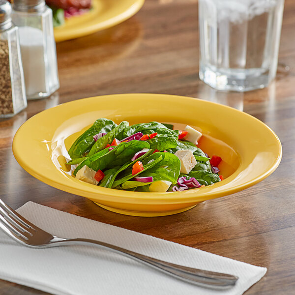 A yellow Acopa melamine salad bowl filled with a salad on a table with a fork and glass of water.