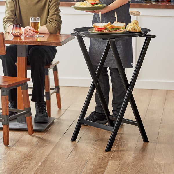A woman using a Lancaster Table & Seating black wood folding tray stand to serve food at a table.