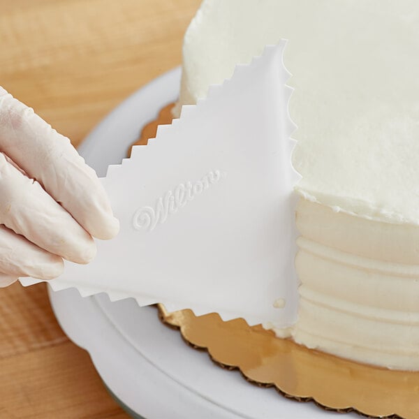 A person using a Wilton plastic triangle icing comb to decorate a white cake.
