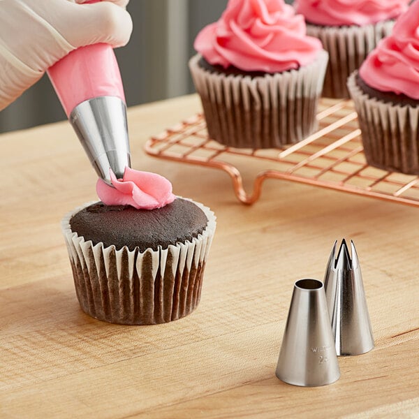 A hand using a Wilton pastry bag with a silver tip to pipe pink frosting on a cupcake.