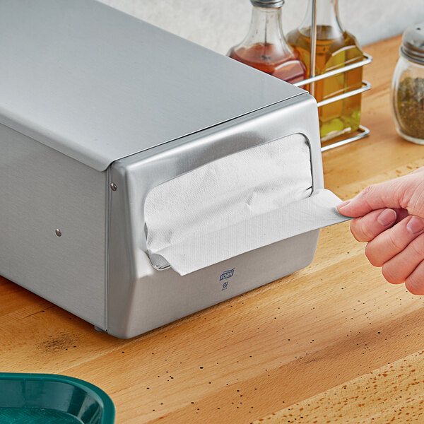 A hand pulling a white Tork dispenser napkin out of a silver box.