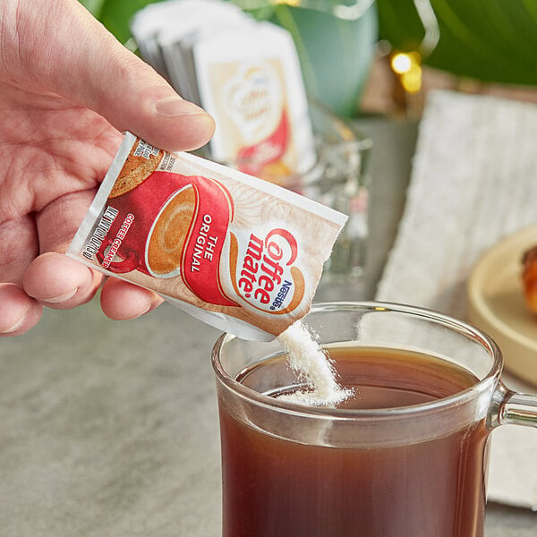 A hand holding a Nestle Coffee-Mate Original single serve coffee creamer packet over a cup of brown liquid.