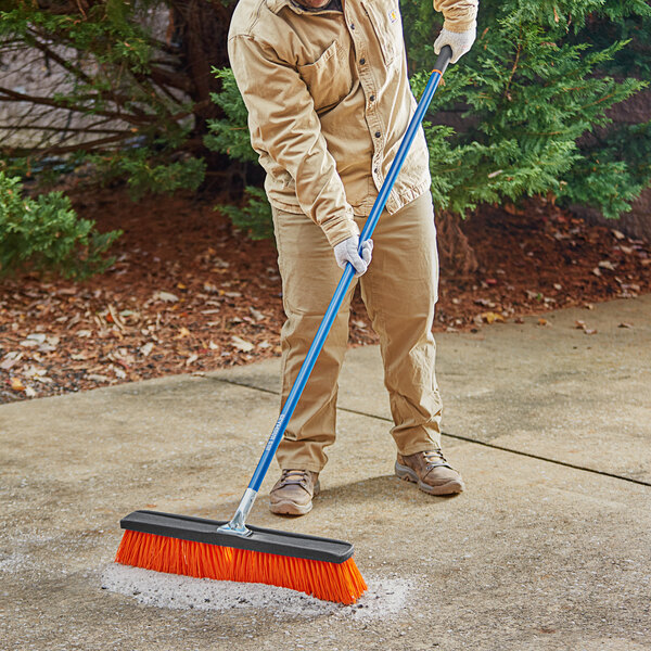 A person using a Seymour Jobsite rough surface street push broom with a blue handle to sweep a sidewalk.