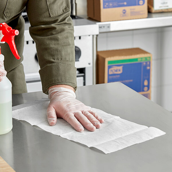 A person wearing clear plastic gloves uses a white Tork heavy-duty cleaning cloth to wipe a hospital cafeteria counter.