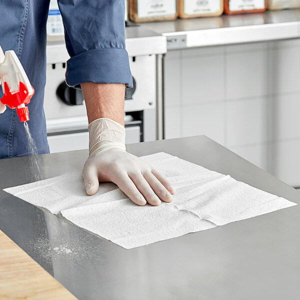 A person in a glove using a white Tork paper wiper to clean a table