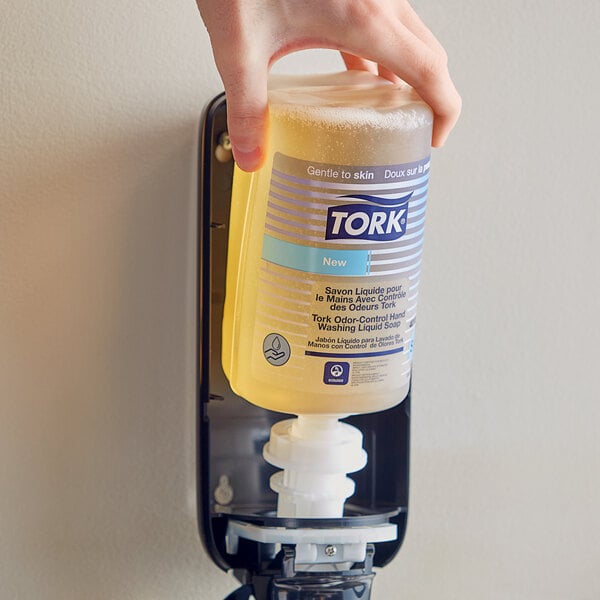 A hand holding a bottle of Tork liquid hand soap in front of a soap dispenser.