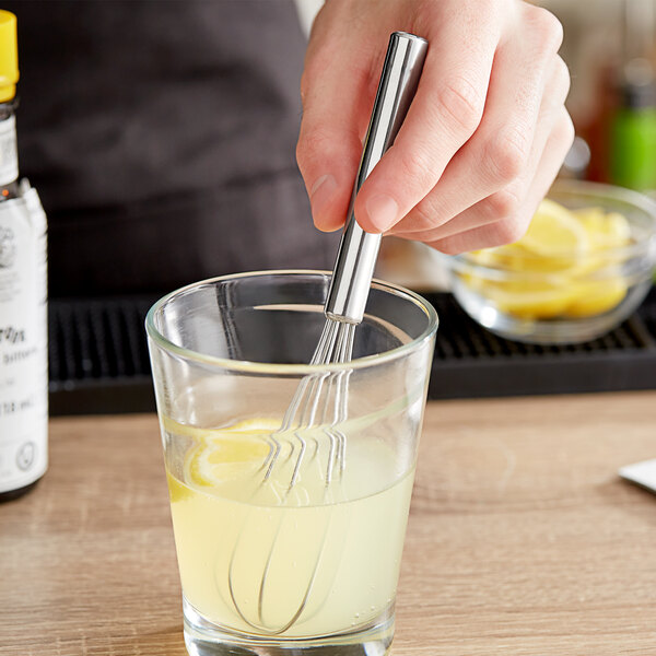 A hand holding a Choice stainless steel mini whisk in a glass of lemon juice.
