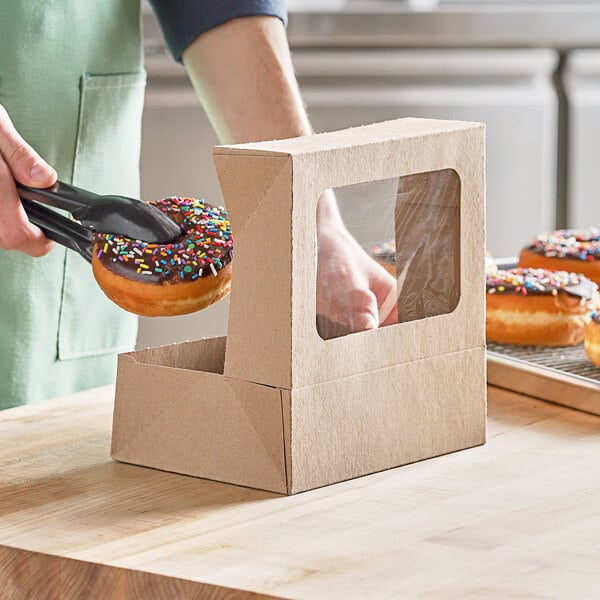 A person holding a chocolate donut with sprinkles in a Kraft window bakery box.