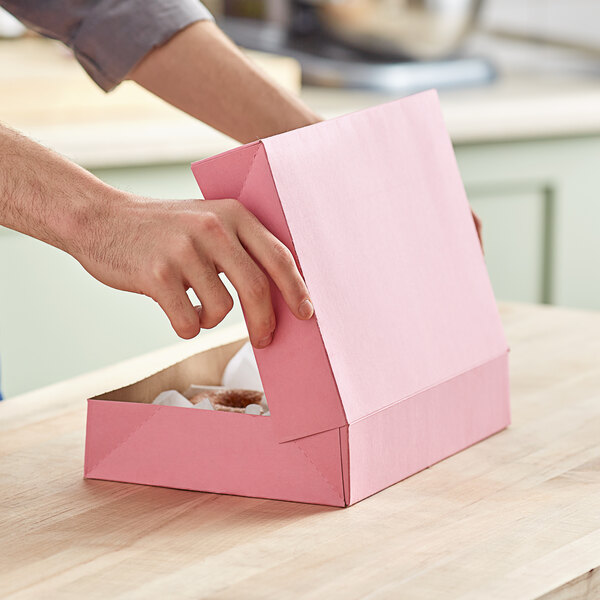 A hand opening a pink Baker's Mark bakery box to reveal a doughnut inside.