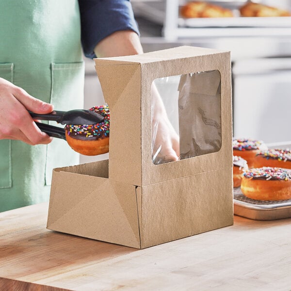 A person using black tongs to pick up a donut with sprinkles from a Baker's Mark kraft bakery box with a window.