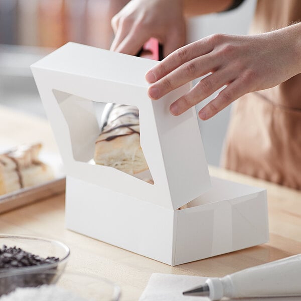 A hand opening a white Baker's Mark bakery box with a plastic window to reveal a piece of cake.