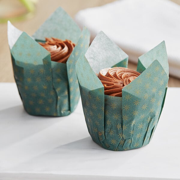 Two cupcakes with chocolate frosting in green Baker's Mark tulip wrappers on a table in a bakery display.