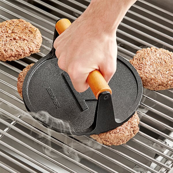 A hand using a Fox Run Cast Iron Flat Grill Press to grill burgers.