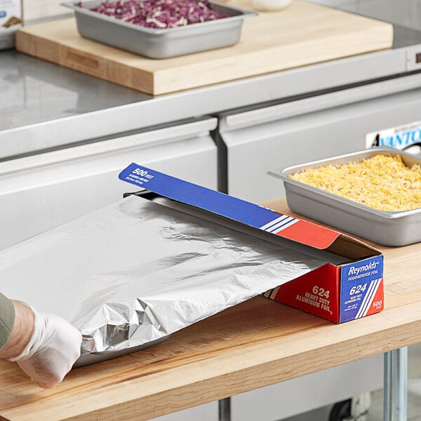 A gloved hand using Reynolds heavy-duty aluminum foil to cover a tray of food.