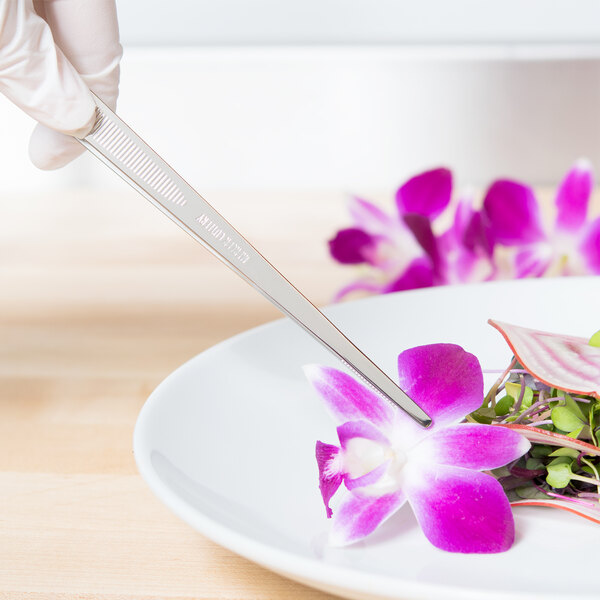 A person's gloved hand using Mercer Culinary straight plating tongs to cut a purple flower on a plate of food.