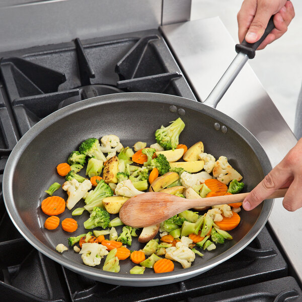 A person cooking vegetables in a Vollrath aluminum non-stick fry pan with a black handle.