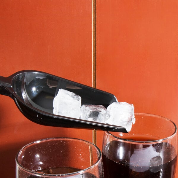A black Fineline utility scoop with ice in it next to a glass of liquid.