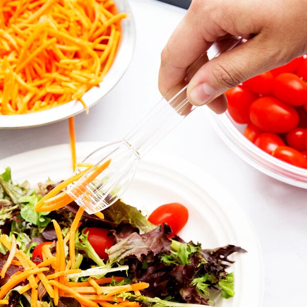 A hand using a Fineline clear plastic tong to serve salad with carrots and tomatoes.