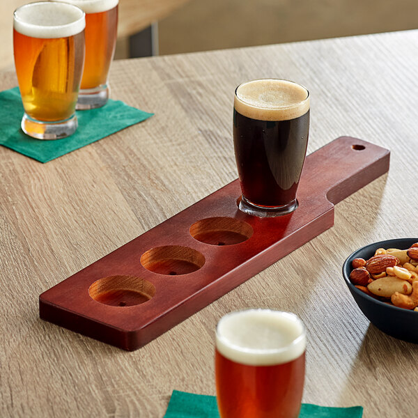 An Acopa mahogany finish flight paddle holding three beer glasses and a bowl of mixed nuts on a table.