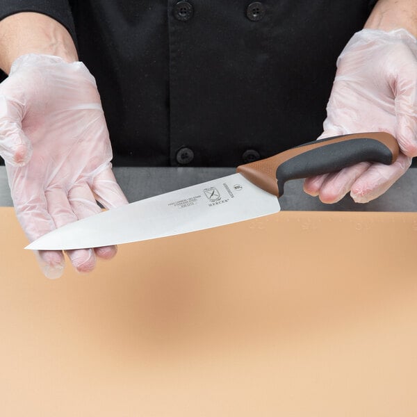 A person holding a Mercer Culinary Millennia Colors chef knife with a brown handle.