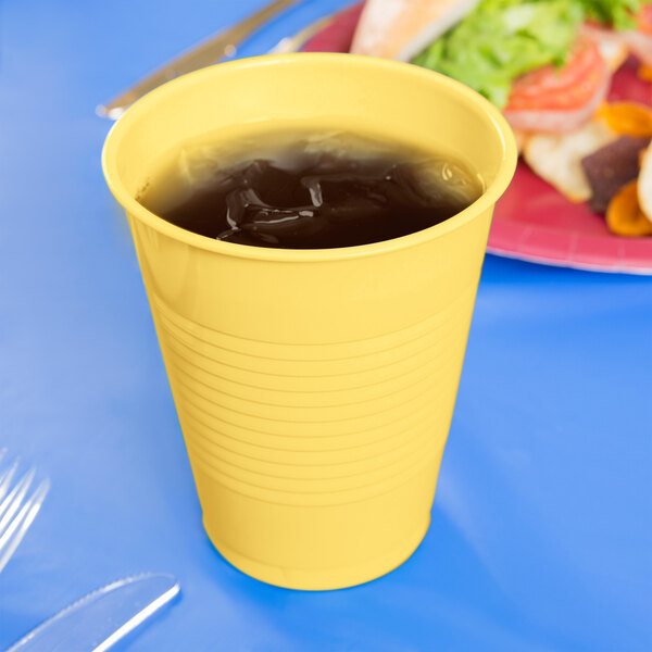 A close up of a yellow Creative Converting plastic cup filled with a yellow drink on a table with food.