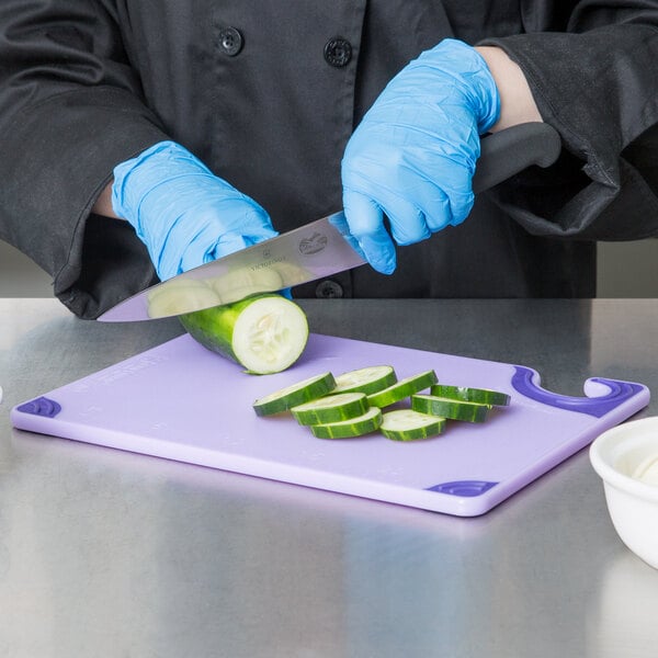 A person wearing blue gloves cutting a cucumber on a San Jamar purple allergen-free cutting board.