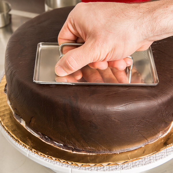 A hand using an Ateco stainless steel fondant smoother to smooth chocolate fondant on a cake.