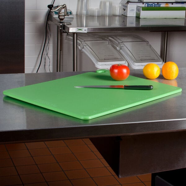A San Jamar green cutting board on a counter with a knife and fruits.