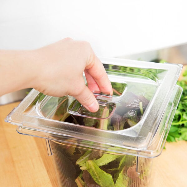 A hand using a Vollrath Super Pan clear polycarbonate lid to cover a plastic food container filled with lettuce.