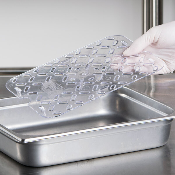 A gloved hand holding a Vollrath Super Pan clear polycarbonate drain tray with holes.