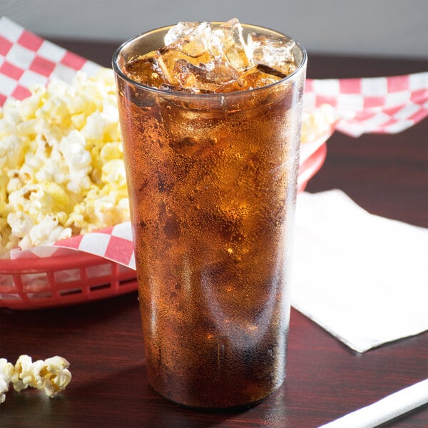 A Cambro amber plastic tumbler filled with soda and ice on a table with a basket of popcorn.