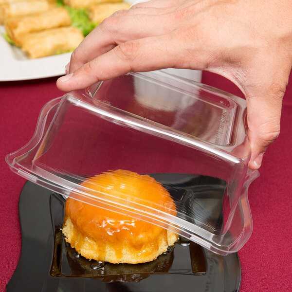 A hand holding a Fineline clear plastic dome lid over a dessert.